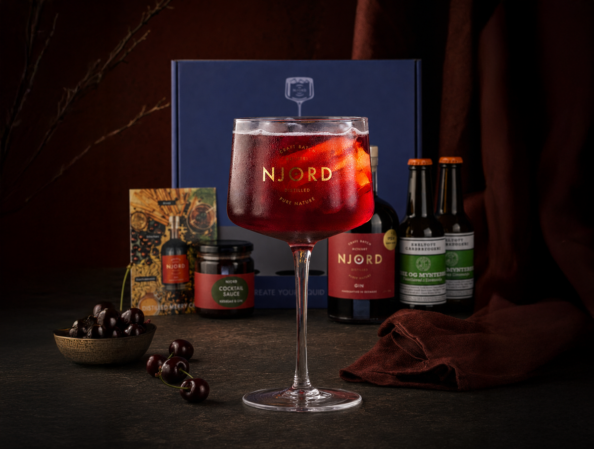 A styled product shot of a NJORD cocktail glass filled with a red cherry cocktail, set against a dark background with a blue NJORD box, a liqueur bottle, two mint and lime sodas, a small jar, a postcard, and cherries on a dark surface.